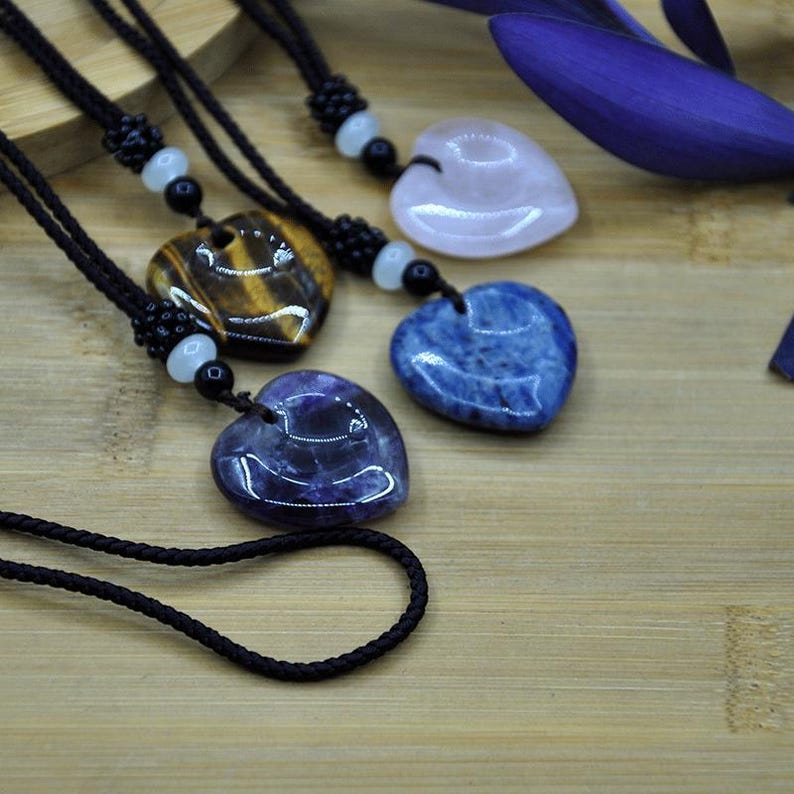 May include: Heart-shaped gemstone pendants on black cords. The pendants are in various colors, including tiger's eye, pink, blue, and purple. Each pendant is strung on a black cord with decorative beads. The background is a light brown wooden surface.