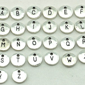 K&ouml;nnte beinhalten: Ein Satz mit 26 silberfarbenen Metall-Anh&auml;ngern mit Buchstaben des Alphabets. Jeder Anh&auml;nger ist rund und tr&auml;gt einen einzelnen Buchstaben des Alphabets in Schwarz.