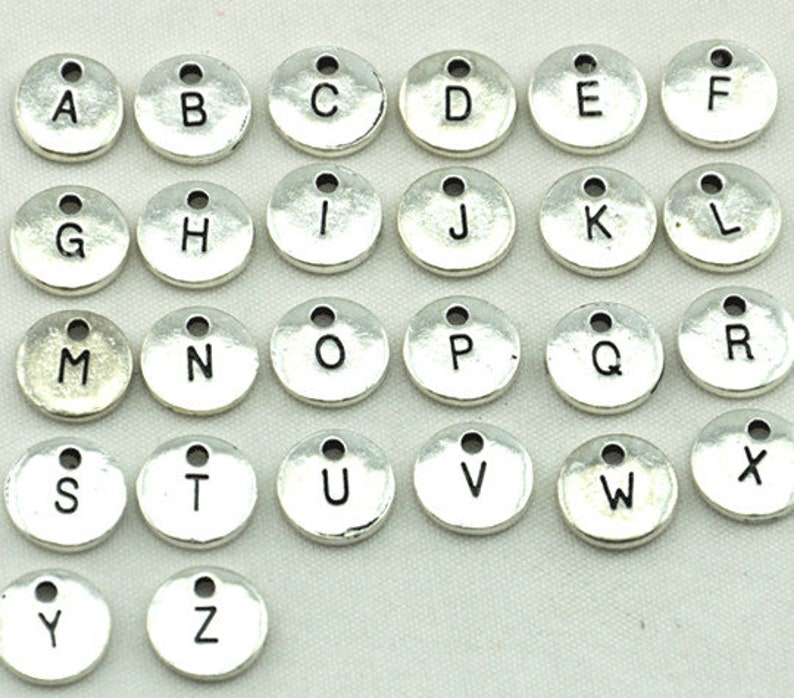 K&ouml;nnte beinhalten: Ein Satz mit 26 silberfarbenen Metall-Anh&auml;ngern mit Buchstaben des Alphabets. Jeder Anh&auml;nger ist rund und tr&auml;gt einen einzelnen Buchstaben des Alphabets in Schwarz.