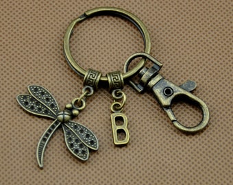 Puede incluir: Llavero de color bronce antiguo con un dije de libélula y un colgante con la letra "B". El llavero incluye un llavero de anilla, un cierre de mosquetón y detalles decorativos. La libélula tiene patrones de alas detallados.