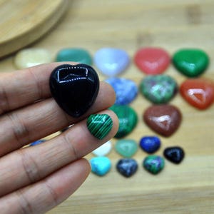 May include: A collection of heart-shaped polished stones in various colors, including black, green, red, and blue. The stones are arranged on a wooden surface.