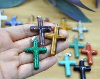 May include: A selection of gemstone crosses in various colours, including green, brown, yellow, and blue. Each cross has a silver-coloured bail for hanging. The crosses are displayed on a light brown surface.