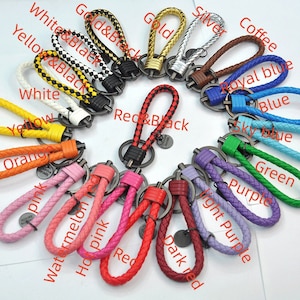 Op de afbeelding: Assortiment van 15 sleutelhangers met gevlochten leren lussen in verschillende kleuren, waaronder rood, roze, oranje, geel, groen, blauw, paars, zwart, wit en bruin. Elke sleutelhanger heeft een metalen ring en een klein metalen label met het woord "Creative" erop.