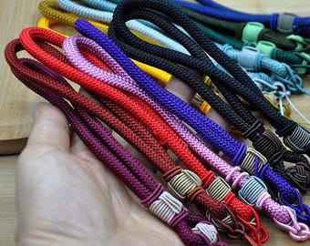 May include: A hand holding a collection of colorful wrist straps. The straps are made of braided cord in various colors, including red, purple, black, and blue. Each strap has decorative knots and tassels.