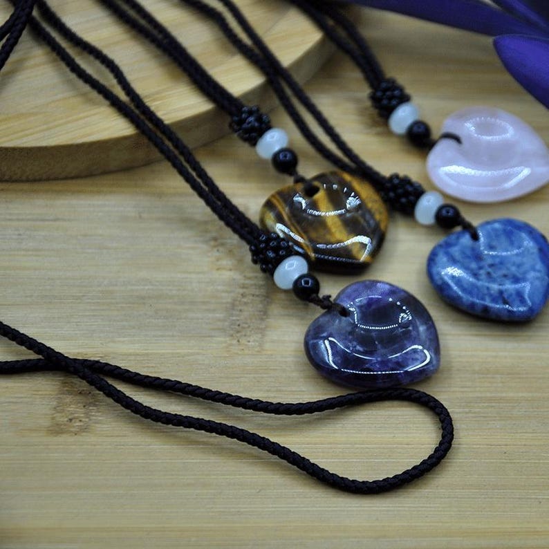 May include: Heart-shaped gemstone pendants on black cords. The pendants are in various colors, including tiger's eye, purple, blue, and pink. Each necklace features a beaded accent near the pendant. The background is a light-colored wooden surface.