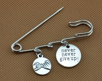 Pode incluir: Alfinete de segurança prateado com dois pingentes. Um pingente tem as palavras "never never give up!" e o outro pingente tem duas mãos segurando os mindinhos.