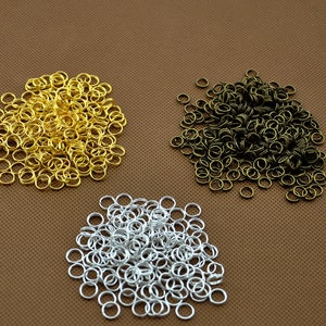 May include: Three piles of metal jump rings in gold, silver, and bronze tones. The jump rings are small, circular, and appear to be made of metal. The background is a solid brown colour.