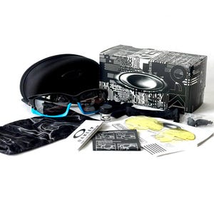May include: Black Oakley sunglasses with blue accents, a black case, and a variety of accessories, including a cleaning cloth, a small bottle of cleaning solution, and a spare lens.