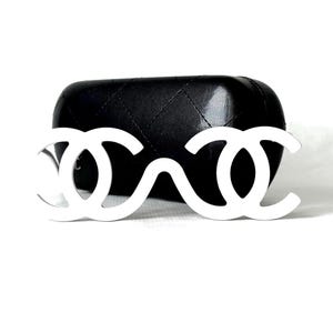 May include: White sunglasses with a distinctive double-C logo, resting in front of a black, quilted eyeglass case. The sunglasses have a unique, open-frame design. The background is white.