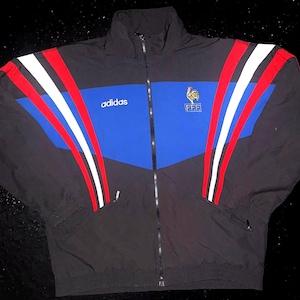 Könnte beinhalten: Eine schwarze, blaue, weiße und rote Adidas-Trainingsjacke mit Reißverschluss und dem F.F.F.-Logo mit Hahn-Emblem auf der Brust. Die Jacke hat einen hohen Kragen, lange Ärmel mit Drei-Streifen-Design und einen durchgehenden Frontreißverschluss.