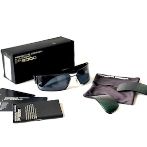 May include: Black sunglasses with a matching case, extra lenses, and a cleaning cloth. The sunglasses have a sleek, modern design with dark lenses and a black frame. The case and paperwork are also black.