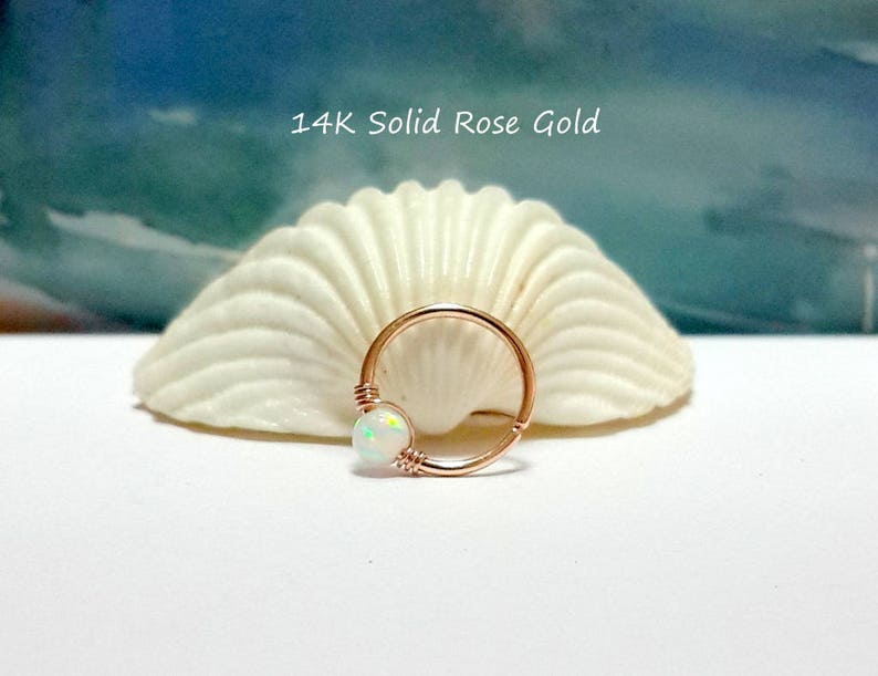 May include: A 14K solid rose gold hoop earring with a white opal gemstone. The earring is on a white seashell background.
