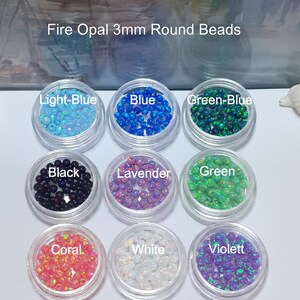 May include: Assortment of 3mm round fire opal beads in various colors, including light blue, blue, green-blue, black, lavender, green, coral, white, and violet. The beads are displayed in small clear containers.