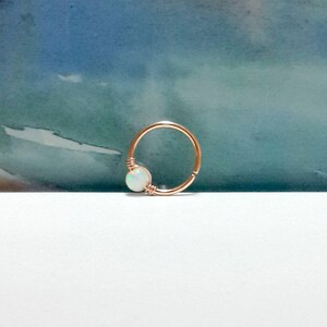 May include: A rose gold hoop earring with a white opal gemstone. The hoop is made of thin metal and the opal is set in a bezel setting.