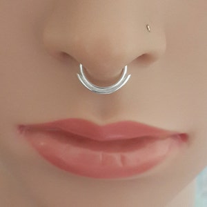 May include: A silver septum ring with a double hoop design. The ring is worn in a person's nose.