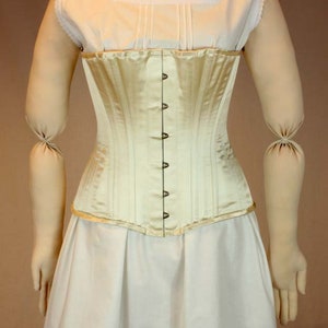 May include: A white satin corset with a row of metal hooks and eyes down the front. The corset is worn over a white cotton chemise with lace trim.