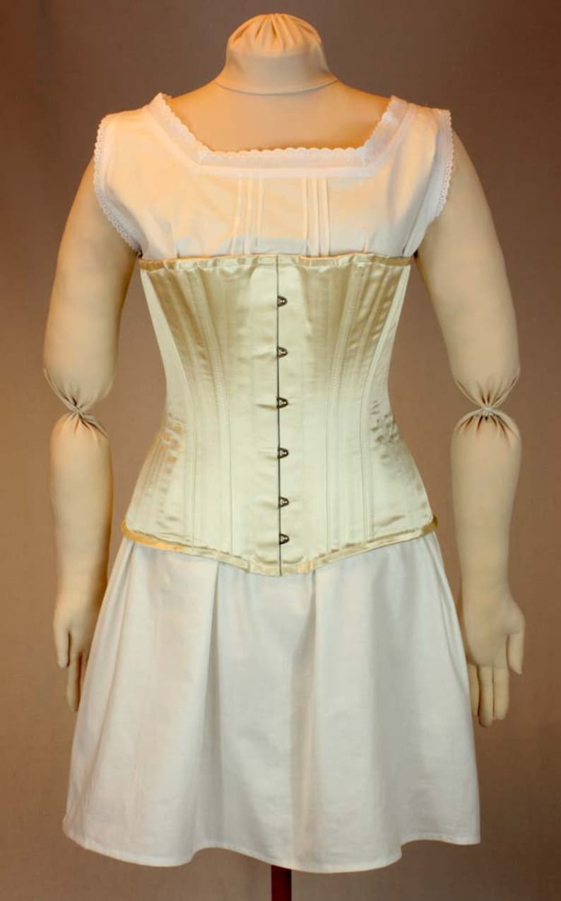 May include: A white satin corset with a row of metal hooks and eyes down the front. The corset is worn over a white cotton chemise with lace trim.