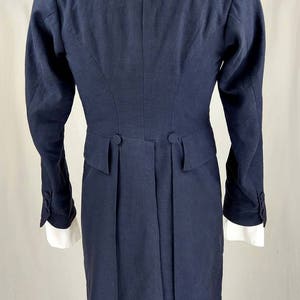 May include: A navy blue tailcoat with a long, fitted silhouette. The coat has a high collar, long sleeves, and a single-breasted closure. The back of the coat features a center vent and two buttons at the waist.