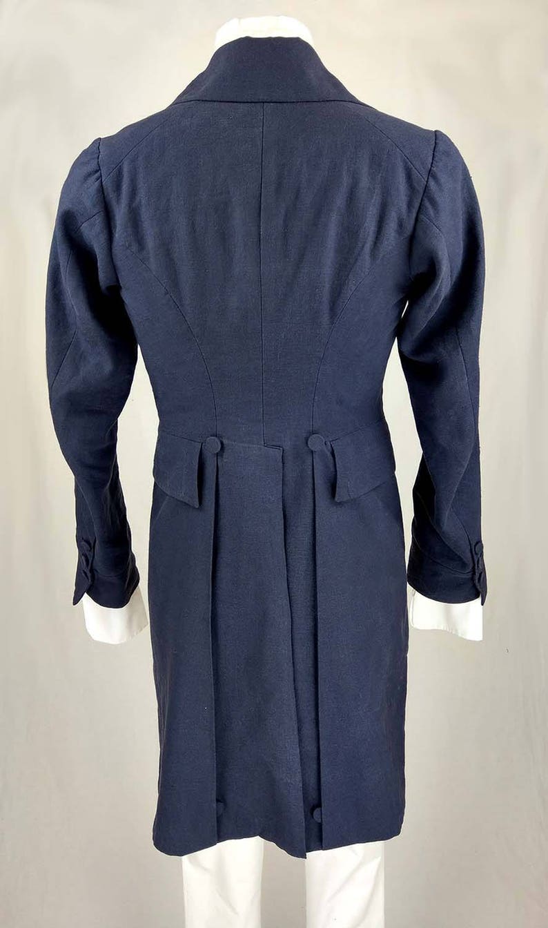 May include: A navy blue tailcoat with a long, fitted silhouette. The coat has a high collar, long sleeves, and a single-breasted closure. The back of the coat features a center vent and two buttons at the waist.