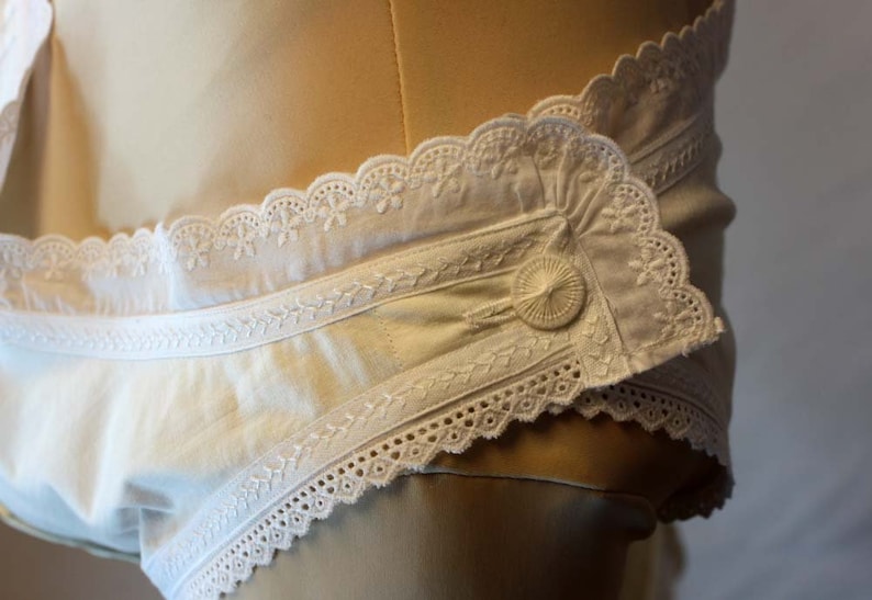 May include: White cotton undergarment with a lace trim and a button fastening. The garment features a delicate embroidered design.