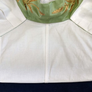 May include: A white cotton garment with a green satin collar featuring gold embroidery. The collar is folded down and the garment is lying flat.