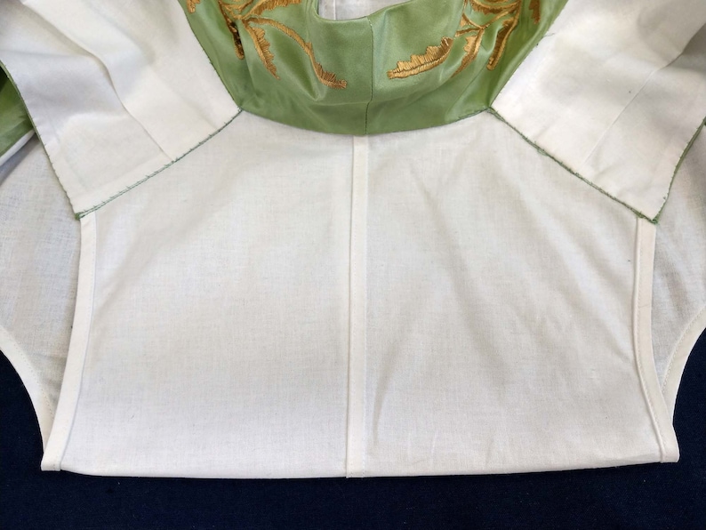 May include: A white cotton garment with a green satin collar featuring gold embroidery. The collar is folded down and the garment is lying flat.
