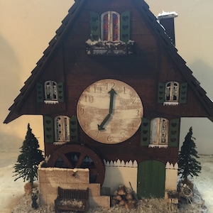 May include: A wooden chalet-style clock with a white face and black hands. The clock has a green door and windows with shutters. The clock is decorated with snow and a small water wheel.