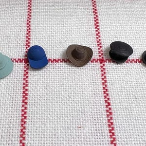 Può includere: Cinque oggetti a forma di cappello in miniatura in vari colori: verde chiaro, blu, marrone e due neri. I cappelli sono disposti in fila su un tessuto bianco a righe rosse.