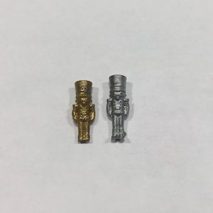 Könnte beinhalten: Zwei Miniatur-Nussknacker-Figuren. Eine ist goldfarben, die andere silberfarben. Beide stehen aufrecht, mit detaillierten Uniformen und Hüten. Die Figuren sind wahrscheinlich aus Metall und werden vor einem schlichten weißen Hintergrund präsentiert.