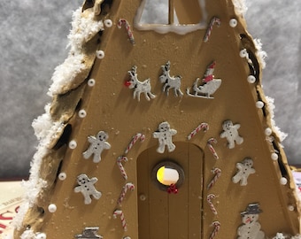 Può includere: Una casetta di pan di zenzero con una finestra ghiacciata bianca e una porta con una ghirlanda. La casa è decorata con bastoncini di zucchero, omini di pan di zenzero e pupazzi di neve. Ci sono anche piccoli alberi davanti alla casa.