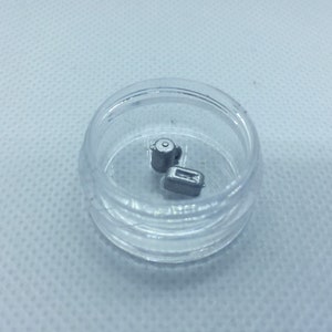 May include: Two small silver metal objects in a clear plastic container. The objects are shaped like a cylinder and a rectangle.