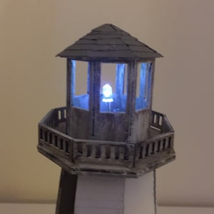 May include: A wooden lighthouse model with a white and black striped base and a brown roof. The lighthouse has a glass lantern with a blue LED light inside.