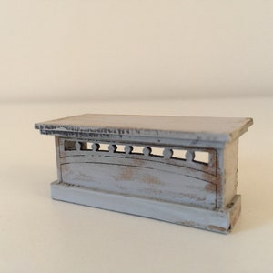 Op de afbeelding: Een miniatuur, lichtgrijze houten tafel met een distressed look, met een decoratief voorpaneel met boogvormige uitsparingen en kleine cirkelvormige accenten. De tafel heeft een rechthoekig blad en een licht verweerde uitstraling, wat wijst op een vintage of rustieke stijl.