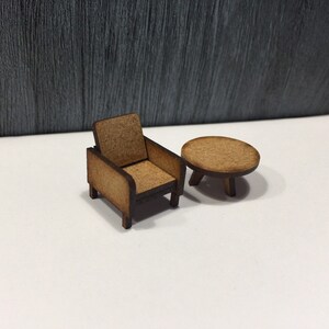 May include: A miniature wooden armchair and a round table, both in a light brown color. The chair has a simple design with a slightly curved back and a square seat. The table has three legs and a flat top.