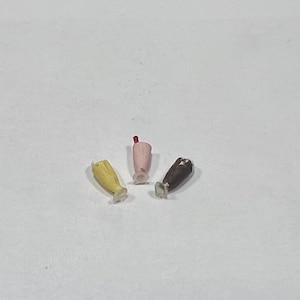 May include: Three miniature milkshakes in yellow, pink, and brown. Each milkshake has a white base and a different coloured top, with a red straw in the pink milkshake. The milkshakes are set against a white background.