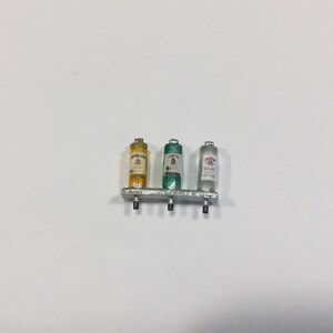 Puede incluir: Tres botellas de licor en miniatura en un estante de metal plateado. Las botellas son de vidrio transparente con etiquetas. La botella de la izquierda es amarilla, la del medio es verde y la de la derecha es transparente con una etiqueta rosa.