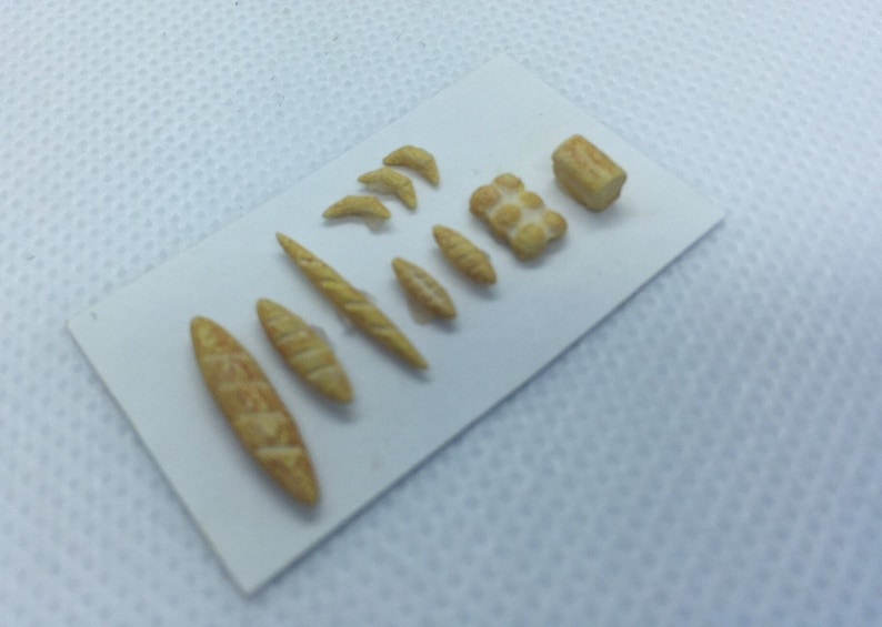 May include: A collection of miniature bread and pastry items, including baguettes, croissants, and a loaf of bread. The items are made of a light brown material and are arranged on a white surface.