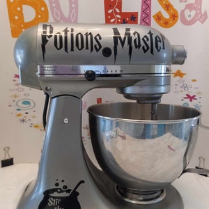Puede incluir: Una batidora de pie plateada con una calcomanía negra que dice "Potions Master" y un diseño de rayo. La batidora tiene un tazón plateado y una calcomanía negra en el lateral que dice "Stir the pot" con un caldero y burbujas.
