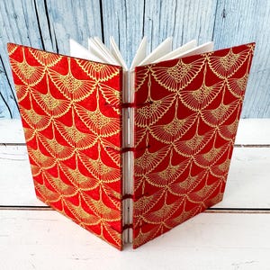 May include: An open journal with red covers adorned with a gold crane design. The pages are white, and the binding is red. The journal is displayed open, revealing the interior pages and the patterned cover.