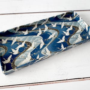 May include: Rectangular decorative tray with a Japanese-inspired design. The tray features a navy blue background with white cranes in flight, gold accents, and stylized waves. The tray is made of a sturdy material and is suitable for serving or display.