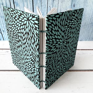 May include: Open journal with teal and black floral patterned covers. The pages are white, and the binding is visible with teal thread. The journal is open, displaying the interior pages against a light blue and white wooden background.