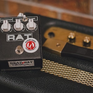May include: A black guitar effects pedal with the word "RAT" in white lettering. The pedal has three knobs labeled "Distortion", "Filter", and "Volume". There is a sticker on the pedal that says "Alchemy Audio".