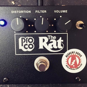May include: Black Pro Co Rat distortion pedal with knobs labeled Distortion, Filter, and Volume. The pedal has a blue LED light and a footswitch. The text "The Rat" is printed in white on the pedal. A sticker for Alchemy Audio is on the bottom right corner.