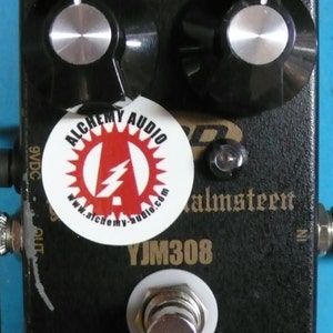 Könnte beinhalten: Schwarzes YJM308 Preamp Overdrive Pedal mit goldener Beschriftung. Das Pedal hat zwei Knöpfe mit der Aufschrift "Gain" und "Level" sowie einen Fußschalter. Ein weißer Aufkleber mit dem Text "Alchemy Audio" und einem roten Blitzlogo befindet sich auf dem Pedal.