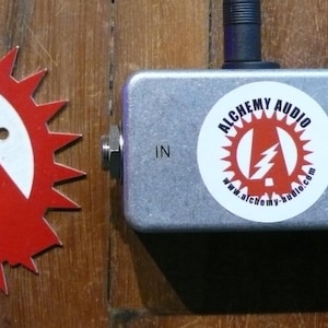May include: A silver metal pedal with a red LED light and the text "Alchemy Audio" with a lightning bolt logo. The pedal has an input and output jack.