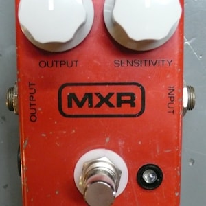 Puede incluir: Un pedal de efectos de guitarra MXR Dyna Comp rojo con dos botones blancos etiquetados "Output" y "Sensitivity", un pedal de pie y conectores de entrada y salida.