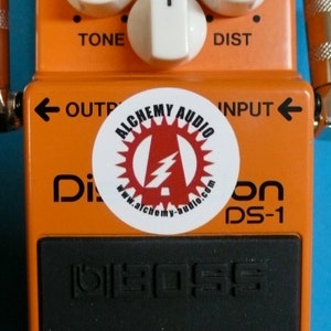 May include: Orange Boss DS-1 Distortion pedal with a white sticker that says "Alchemy Audio" and has a lightning bolt logo. The pedal has three knobs labeled "Tone", "Dist", and "Check". The pedal has an input and output jack and a black "Boss" logo on the front.