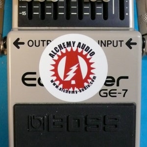 May include: A gray Boss GE-7 Equalizer guitar effects pedal with a black rubber foot switch and a white sticker with the text "Alchemy Audio" and a lightning bolt logo. The pedal has a row of seven knobs with white knobs and black markings.