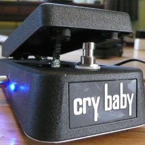 May include: A black Cry Baby wah pedal with a blue LED light. The pedal has a foot switch and a knob for adjusting the wah effect.