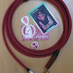 May include: Red guitar cable with a right-angle plug on one end and a straight plug on the other. The cable is wrapped in a red braided sleeve. The cable is coiled up and there are three small stickers and a card with a picture of a musician on it.